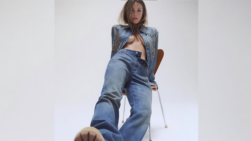 Leila George in denim jeans with an embellished denim jacket, seated on a chair, legs extended toward camera.