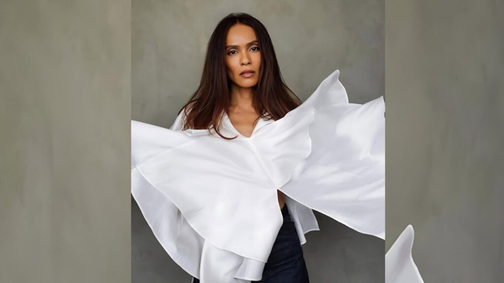 Lesley-Ann Brandt with long dark hair in a white blouse with oversized sculptural sleeves, posing before a gray studio backdrop.