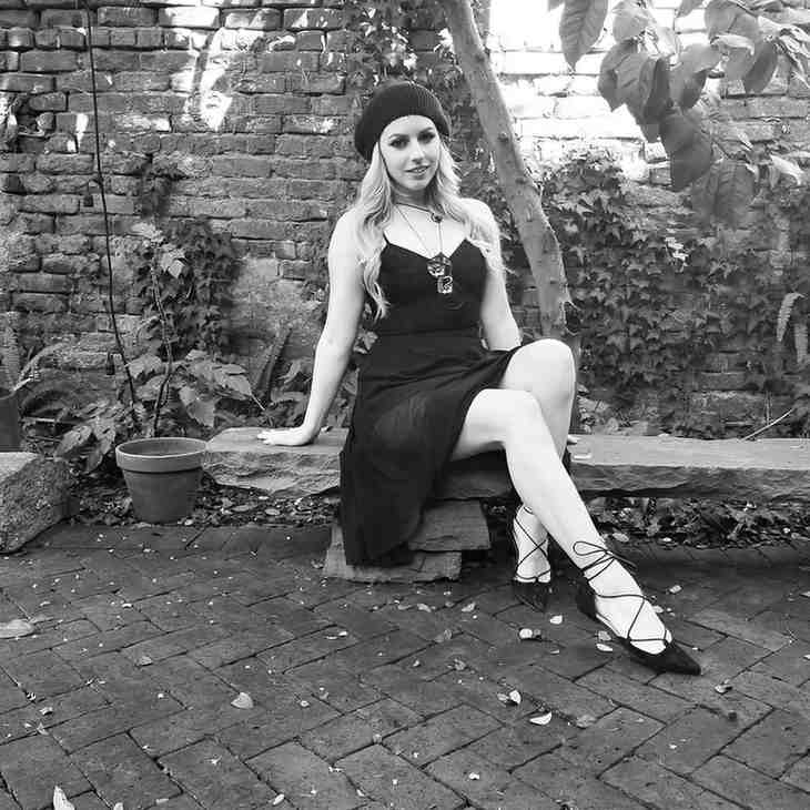 Lexi Belle sitting on a stone bench in a garden, wearing a black dress, beanie, and strappy heels with a pendant.