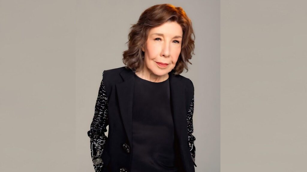 Lily Tomlin with shoulder-length brown hair in a black blazer with sequined sleeves, against beige backdrop.