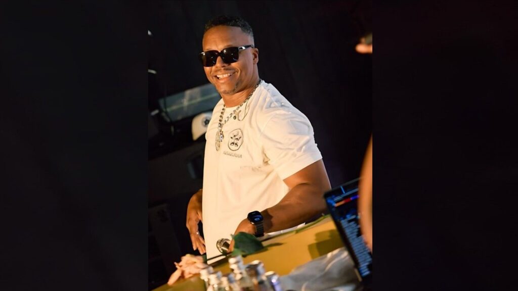 Lupe Fiasco in a white tee and sunglasses stands at a DJ booth, wearing a chunky chain and watch, with a mixer nearby.