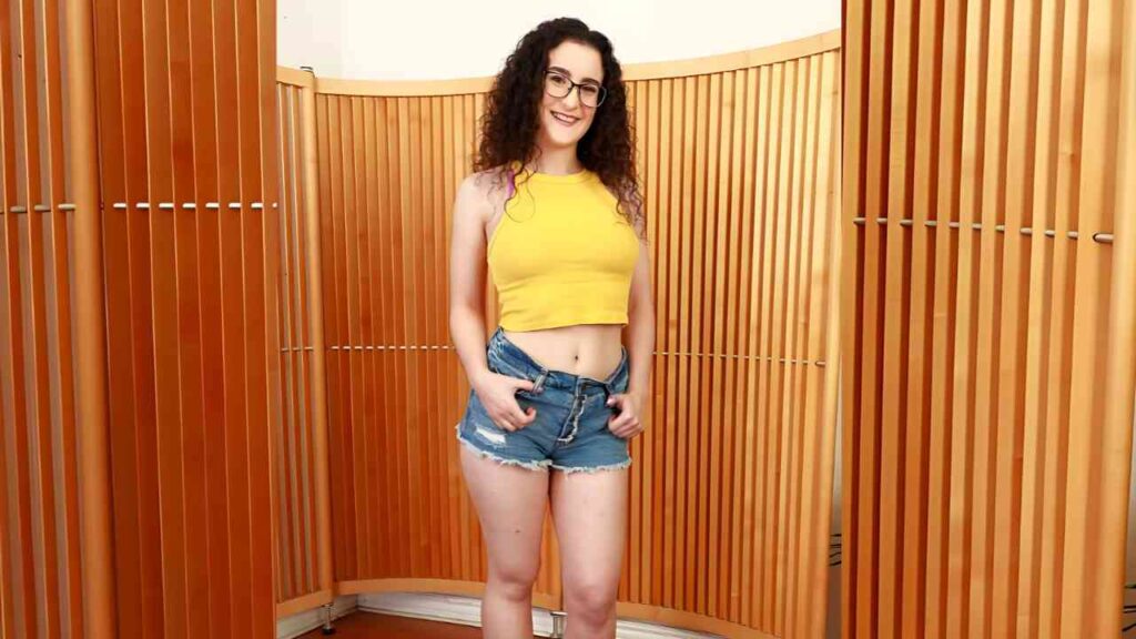 Lyra Lockhart with curly hair and glasses stands in front of warm wooden slat panels, wearing a yellow crop top and denim shorts.
