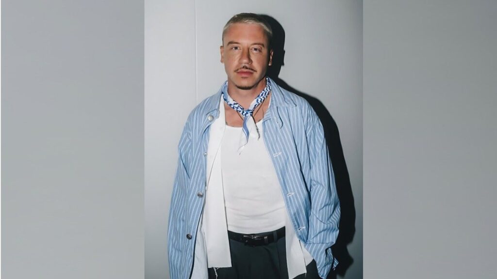 Macklemore with a mustache poses against a gray backdrop, wearing a striped shirt over a white top and a blue scarf.