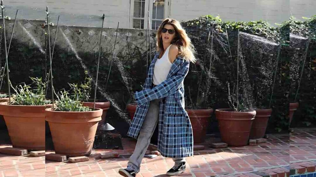 Marisa Tomei in a white tank top, blue plaid coat, jeans, and sunglasses walks along brick patio, with pots and mesh fence behind.