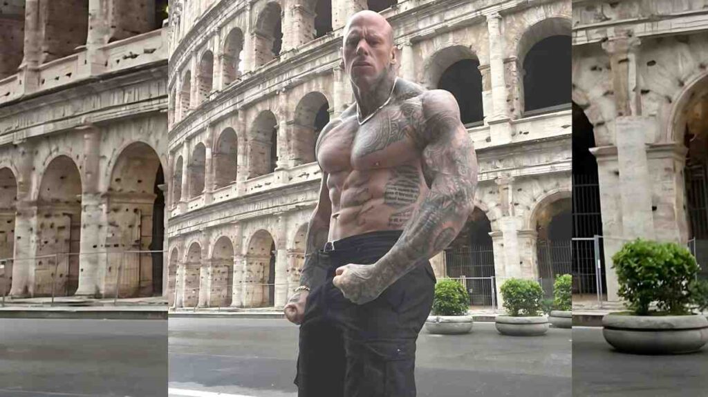 Martyn Ford bodybuilder stands shirtless before ancient Colosseum, fists clenched, in dark cargo pants and a silver chain.