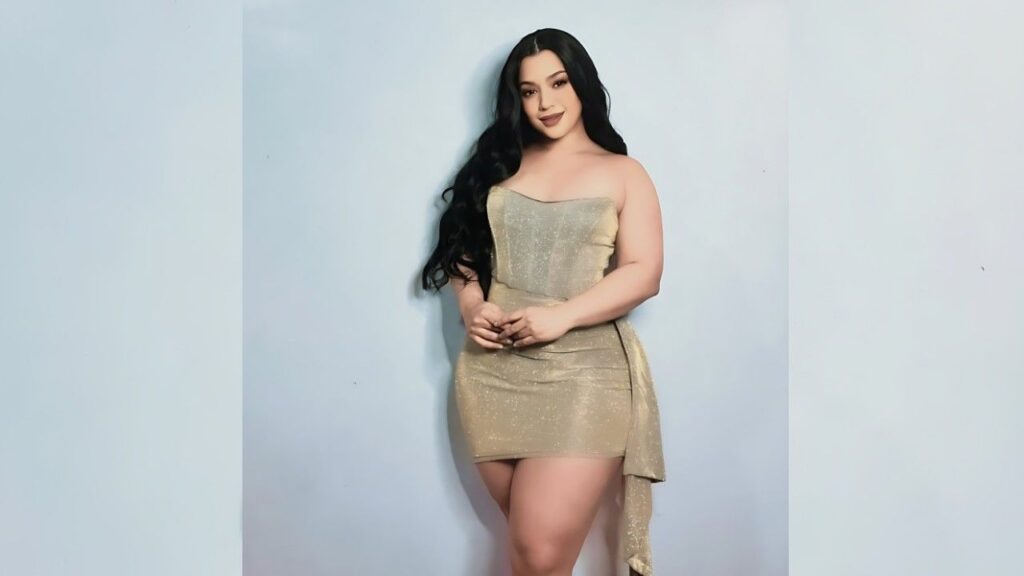 Mimi Curvaceous in a shimmering gold strapless mini dress with draped side fabric, standing against pale blue backdrop.
