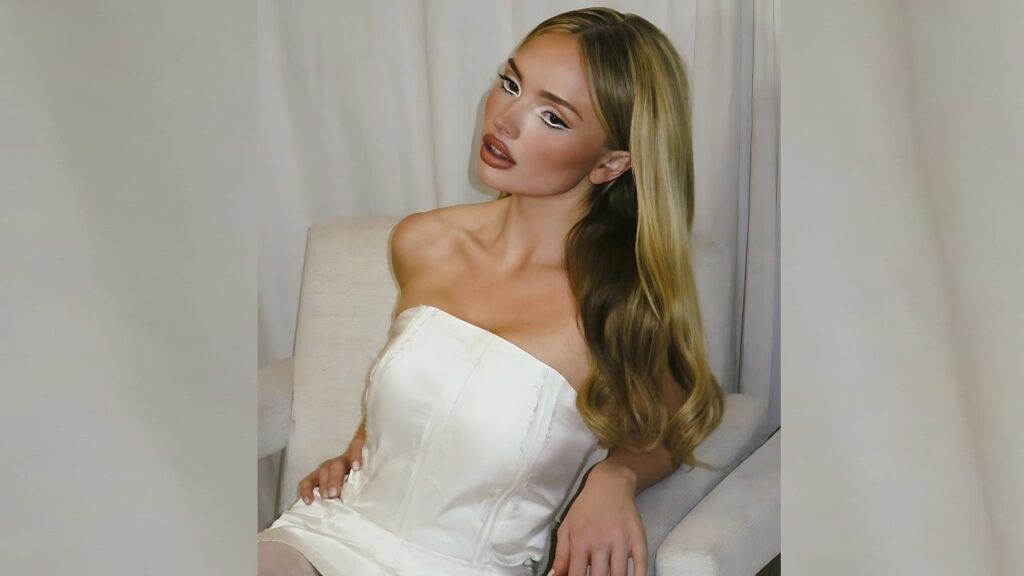 Natalie Alyn Lind with wavy hair sits on a light chair in a white strapless corset top, gazing to the side against curtains.