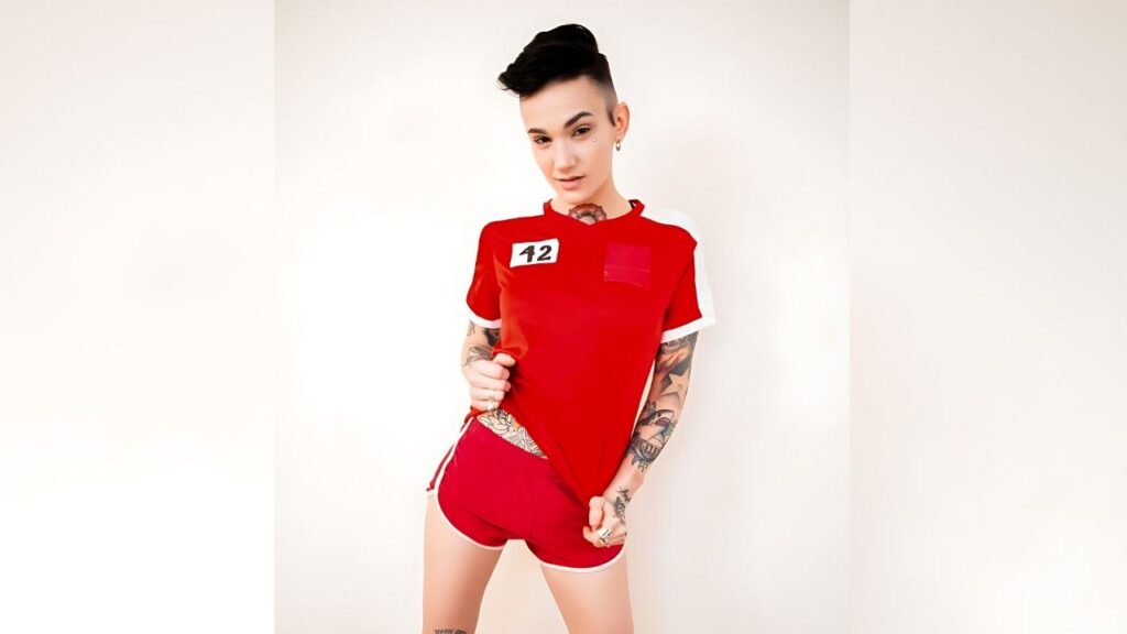 Nikki Hearts with short black hair in a red athletic top and shorts, 42 on chest, tattoos on arms, posing against a pale wall.