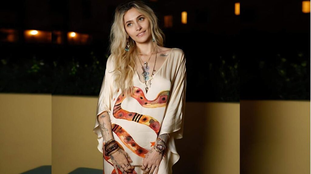 Paris Jackson in a cream flowing dress with orange snake print, layered necklaces and tattoos, amid warm night lights.