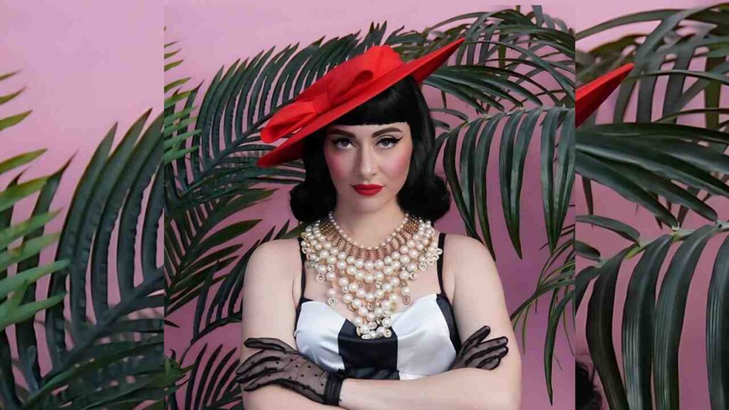 Qveen Herby in a black-and-white satin dress, layered pearls and gloves, red hat, arms crossed against pink backdrop.