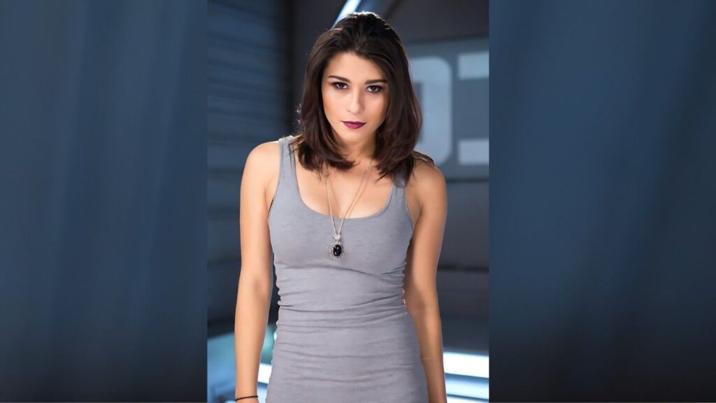 Raven Rockette with shoulder-length dark hair in a gray tank top and a black pendant necklace, gazes at the camera with a subtle smile.