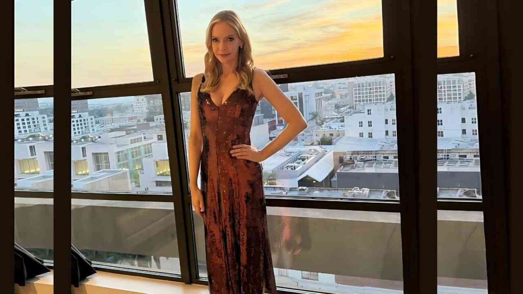 Riki Lindhome in a burgundy sequin floor-length gown stands with one hand on her hip beside large windows at sunset.