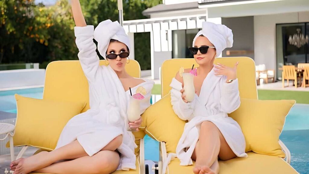 Rosanna Pansino in white bathrobes with towel turbans lounge by a pool on yellow chairs, sipping tropical drinks with umbrellas.