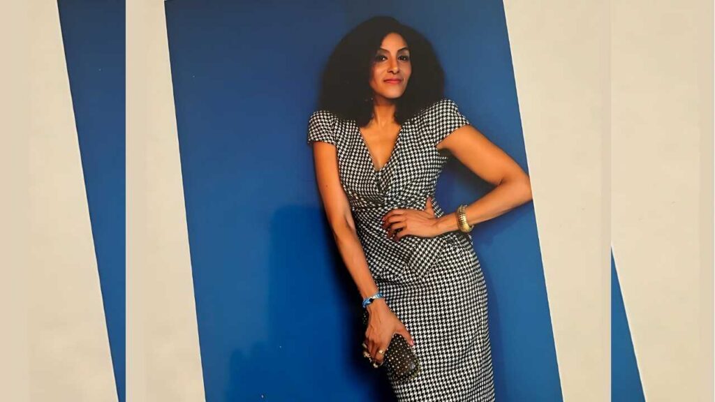 Sarah Jones with voluminous curls poses in a black-and-white houndstooth wrap dress against blue panels, clutch in hand.