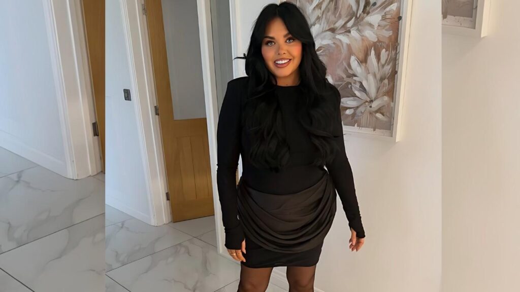 Scarlett Moffatt in a sleek black long-sleeve dress with a draped hip wrap stands in a hallway, marble floor and framed art.