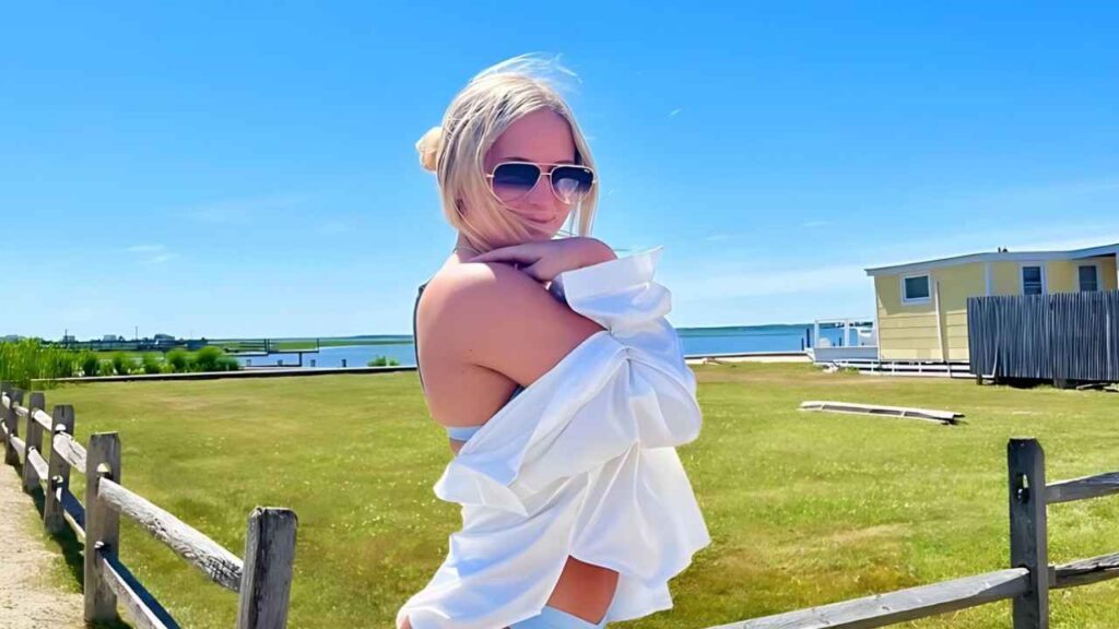 Sierra Nicole in sunglasses stands near a wooden fence on a sunny coast, shirt draped over her shoulders, blue water behind.