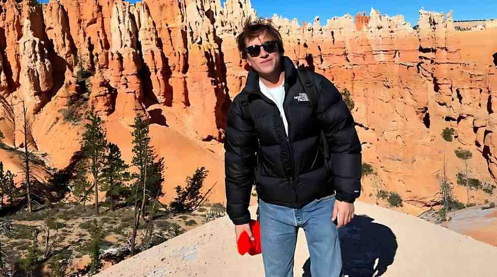 Skyler Gisondo in a black puffer jacket and jeans stands on a sandy overlook with orange cliffs behind him, holding a red item.