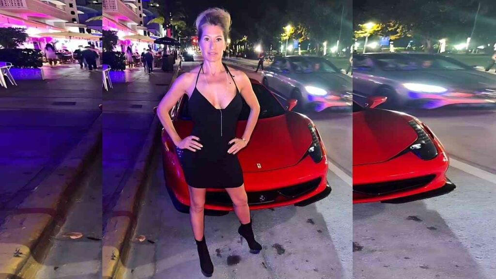 Sloan Rider in a tight black zip-front mini dress stands with hands on hips beside a red Ferrari, on a neon-lit nightlife street with blurred moving cars.