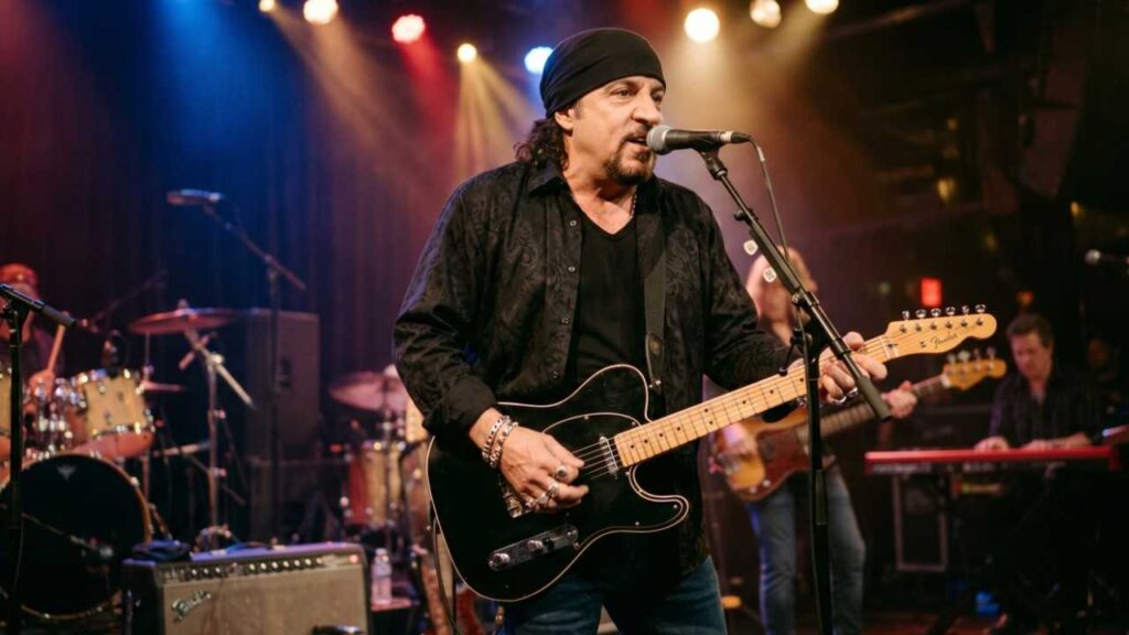 Steven Van Zandt in a black bandana and shirt plays a black Tele-style guitar into a mic on a club stage, with drummer and keyboardist in back.