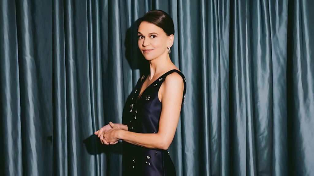 Sutton Foster with pulled-back hair and hoop earrings stands before blue curtains in a navy sequined dress, hands folded.
