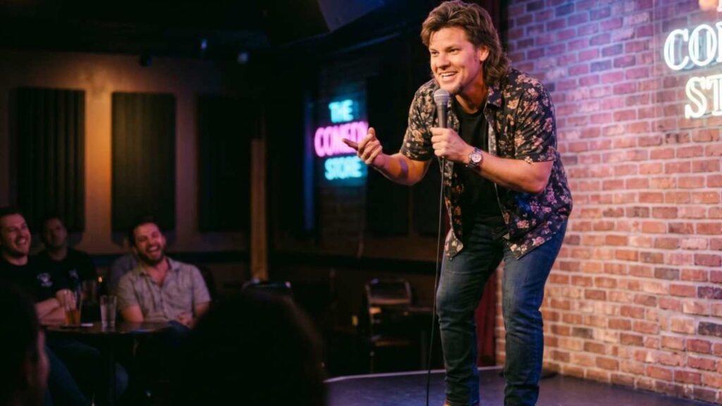 Theo Von in a floral shirt and jeans, leaning into a microphone on a brick stage neon sign behind audience laughing.