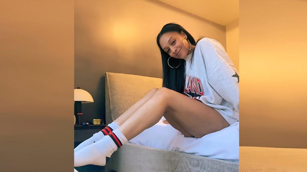 Tia Mowry with long dark hair sits on a hotel bed in a gray oversized sweatshirt, white socks with red stripes, smiling.