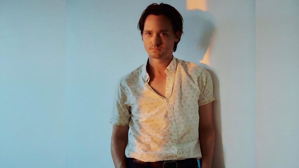 Tom Schilling with dark hair leans against a pale blue wall, wearing a light patterned shirt; warm light from the right.