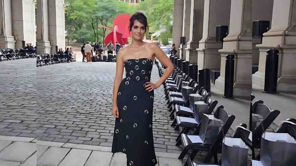 Torrey DeVitto with circular metal accents, she poses with hand on hip under a colonnade beside chairs and bags.