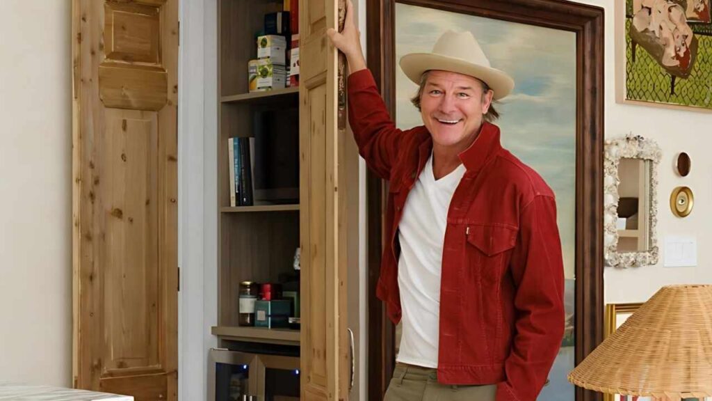 Ty Pennington in a red jacket and beige hat stands by a cabinet with its door ajar; shelves visible, painting behind.