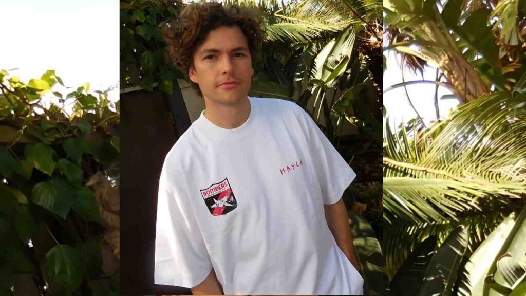Vance Joy with curly brown hair wearing a white oversized tee with a red chest logo and a patch, standing among plants.