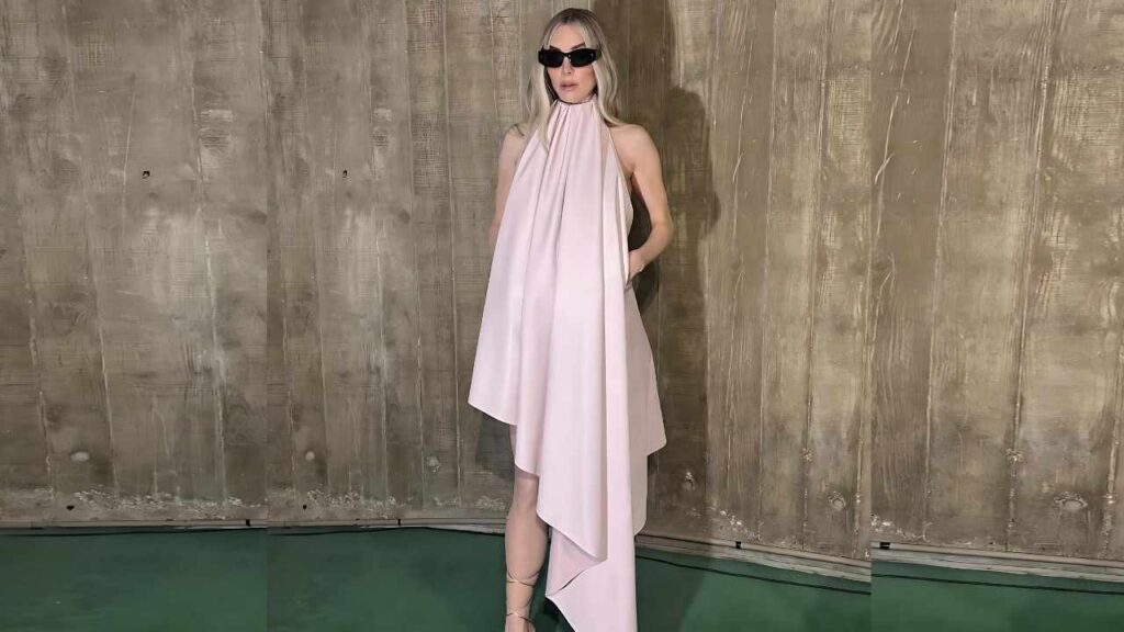 Vanessa Kirby in pale pink draped halter dress with asymmetrical hem, black sunglasses, posed before a weathered wooden wall.