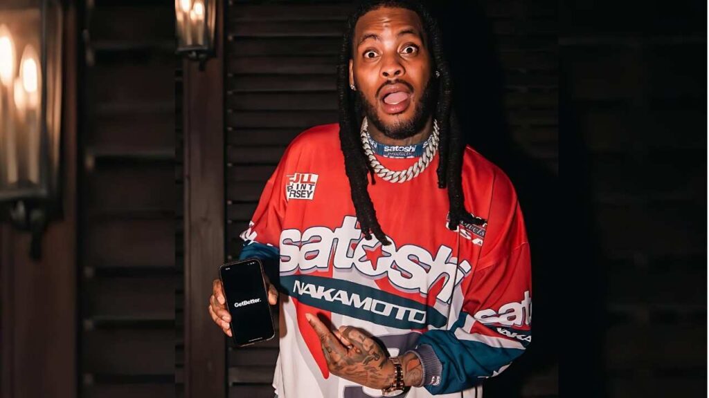 Waka Flocka with braided hair and a chunky chain wearing a red-white-blue jersey holds a phone showing GetBetter.