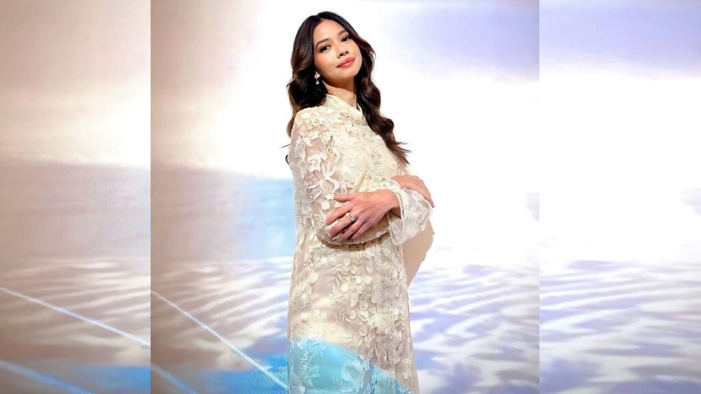 Yuki Kato in a cream lace dress stands against a soft pastel backdrop, arms crossed over her belly with a gentle smile.