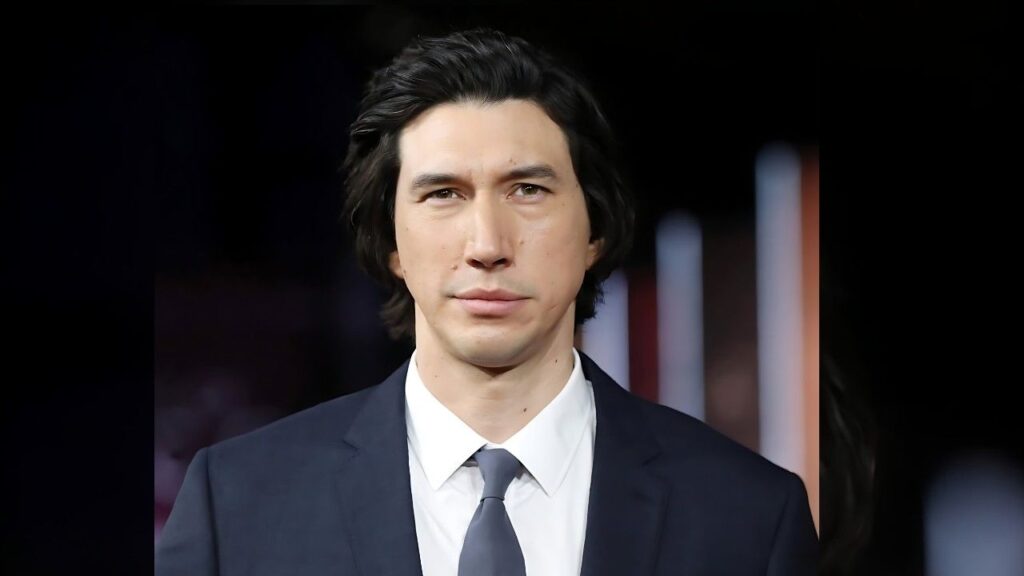 Adam Driver with dark, swept-back hair in a navy suit and light gray tie, against a dark blurred backdrop.