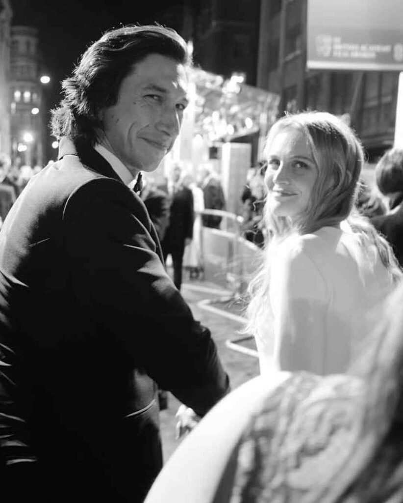 Black-and-white street scene of a stylish couple; Adam Driver in tuxedo and Adam Driver wife in light coat smile at the camera amid city lights.
