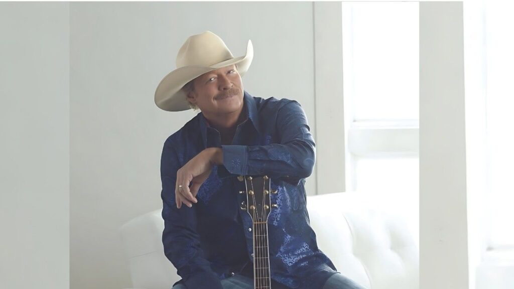 Alan Jackson in a blue shirt sits with a guitar, resting his arm on the neck, in a bright white room.