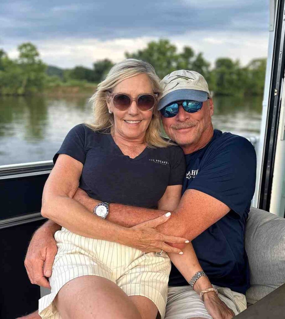 Alan Jackson wife on a boat, arms around each other, wearing sunglasses, with a lake and trees beyond under a cloudy sky.