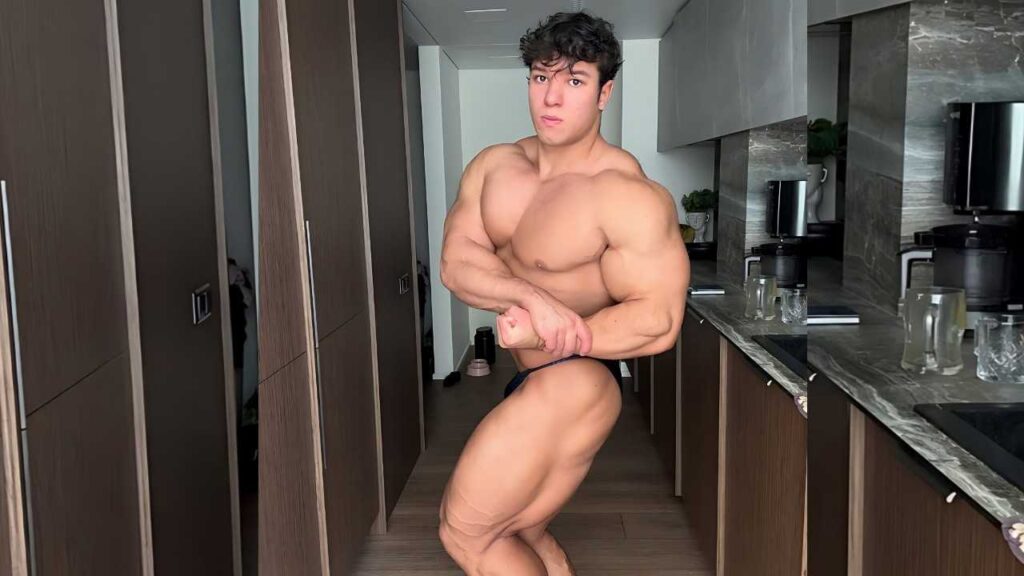 Albertas Krinickis flexes biceps in a modern kitchen hallway, wearing dark briefs amid wood cabinets and marble counters.