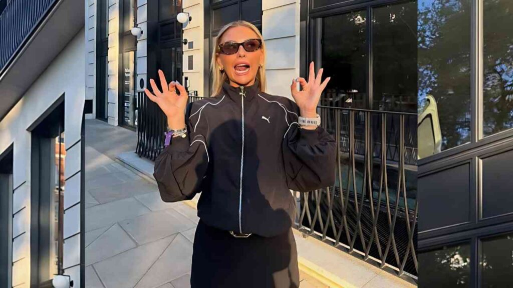 Alysia May McAndrew in a black Puma jacket and sunglasses flashes an OK gesture on a city sidewalk, with a glass building behind.