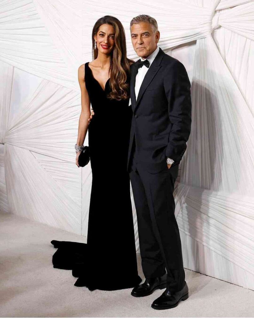 Amal Clooney in a black evening gown beside George Clooney in a classic tuxedo, posing against a white draped backdrop.