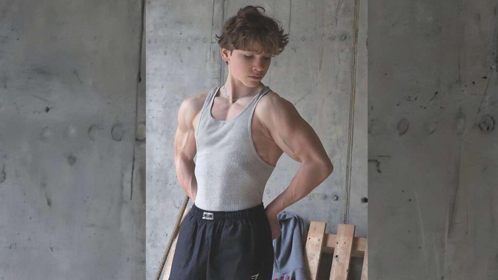 Andy Andrici in a gray tank top and black shorts stands against a concrete wall, hands on hips, looking down.