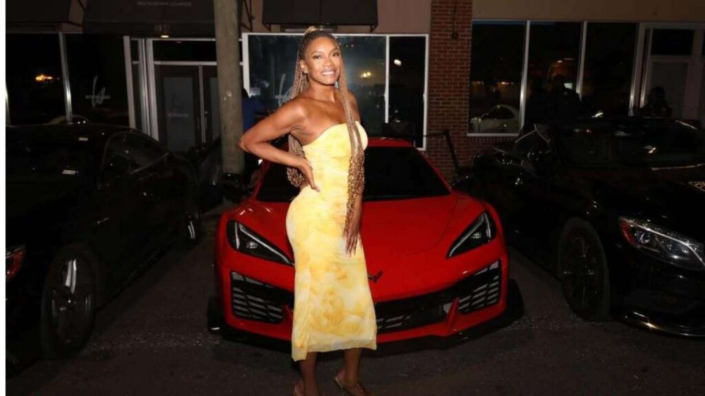 Ashley Neice in a yellow strapless dress with long braids poses with hand on hip in front of a bright red Corvette.