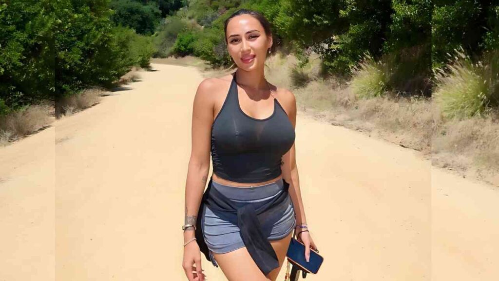 Astrid Nelsia walks a sunlit dirt trail in a cropped tank top and gray shorts, jacket tied at the waist, holding a phone.