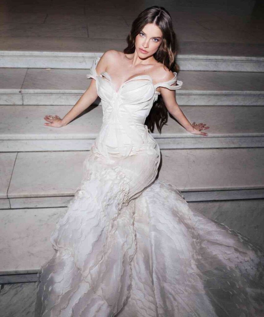 Barbara Palvin in a white off-shoulder corseted gown sits on marble stairs, hands on steps, flowing ruffled train.