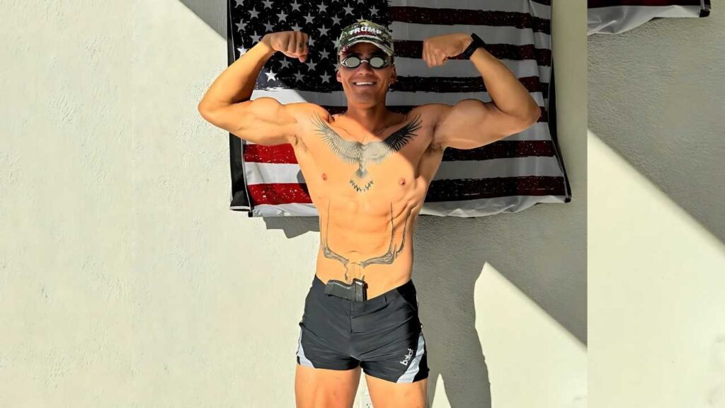 Brandon Banach shirtless in black shorts flexes biceps before flag backdrop; eagle chest tattoo, camo cap with Trump, goggles.