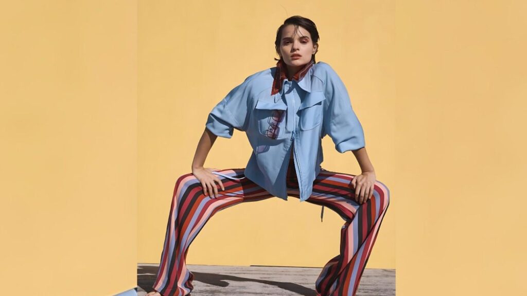 Brianna Hildebrand in a light-blue oversized jacket and striped pants adopts a wide stance against a yellow backdrop, hands on knees.