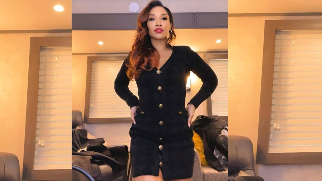 Candice Patton in a black long-sleeve dress with gold buttons stands in a warmly lit room, hands on hips, wavy auburn hair.