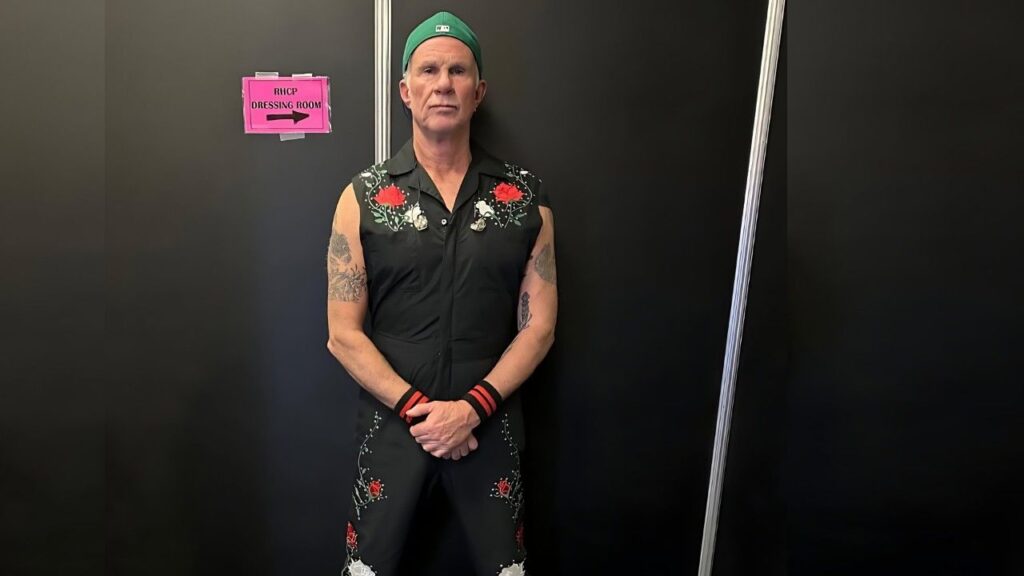 Chad Smith in sleeveless black embroidered outfit and green cap, tattooed arms, stands against a dark wall beside a pink RHCP sign.
