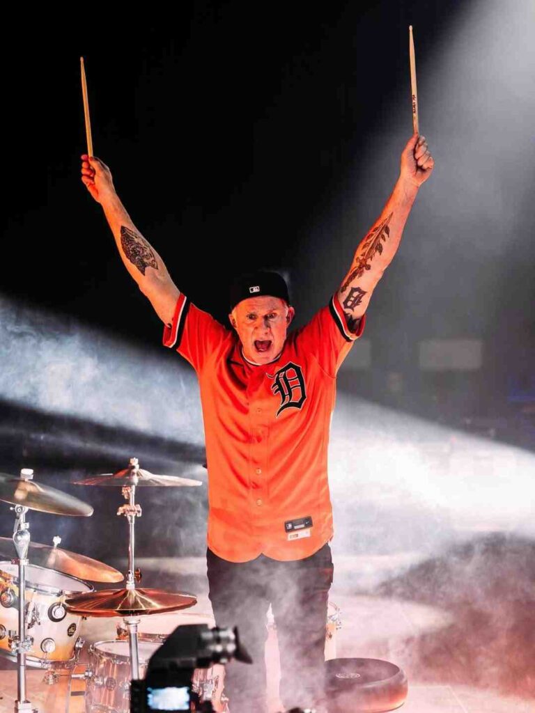 Chad Smith Band on stage raises drumsticks high, wearing an orange jersey with a black D logo, tattoos visible, smoky lights behind.