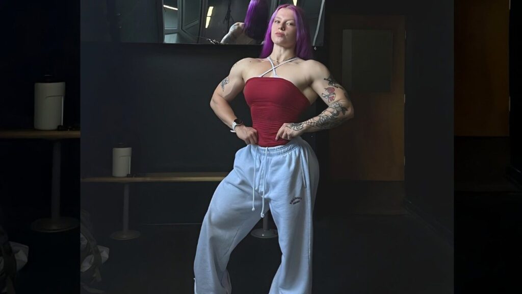 Charlotte Schaech with purple hair flexes in a dim studio, wearing red halter top and gray sweatpants, tattoos visible on arms.